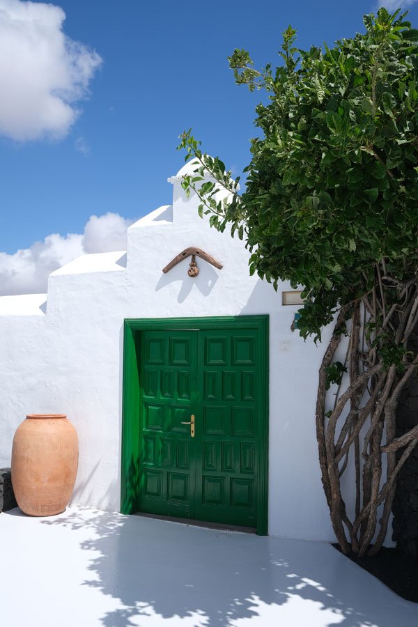 whitewashed building with green door framed by a tree and a large earthenware pot under a blue sky