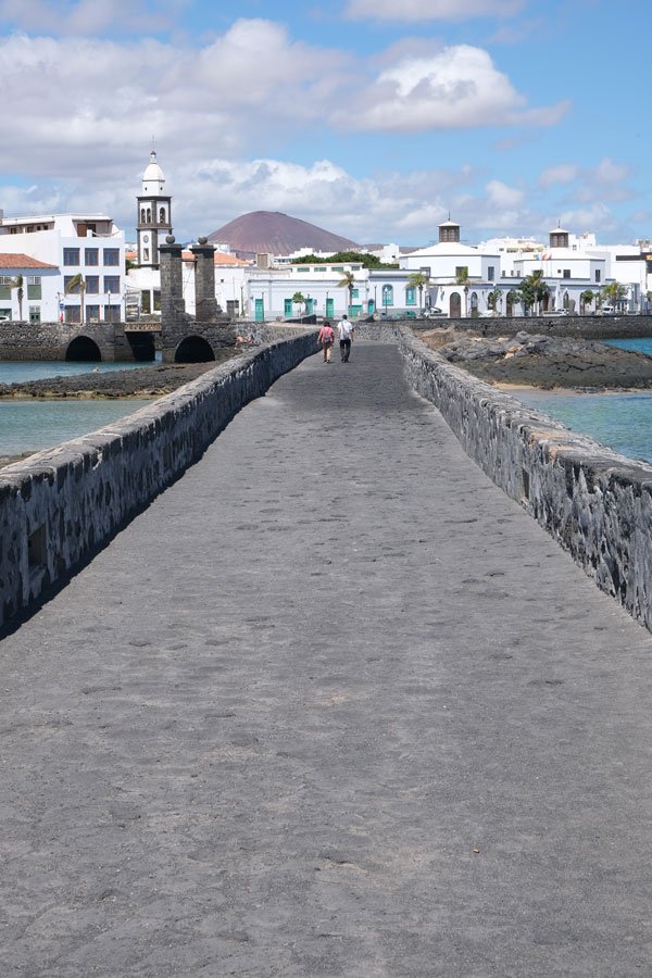 2 people walling alone a stone footbridge twards whitewashed buildings in arrecife lanzarote
