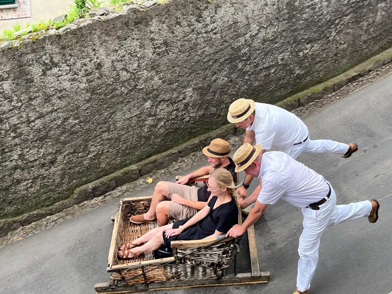 two men dresssed in whote ouching a wicker toboggan at seppd down a hill in monte madeira