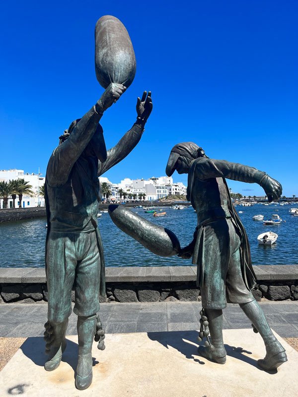 bronze sculpture to 2 people playing a game with oval bats in front of a pretty lagoon