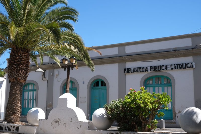 low whiteswashed building with green doow in a small square with a palm tree