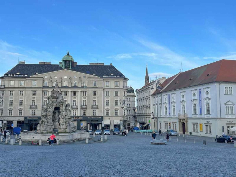 large cobblestone plaza lined with medieval buildings and with a stone fountain in cnetre