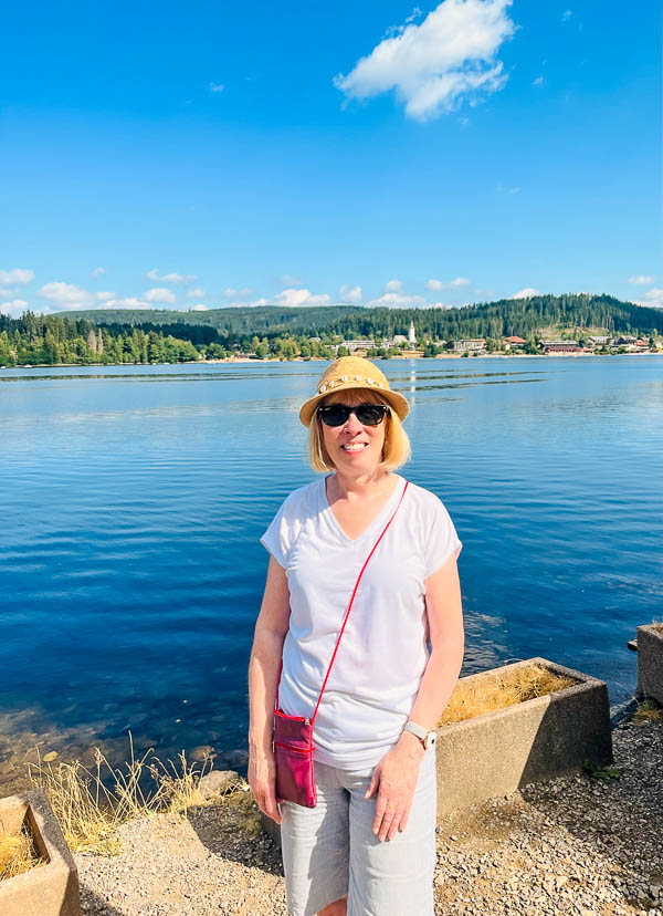 woman in t shirt and shorts and hat standing by the side of a lake